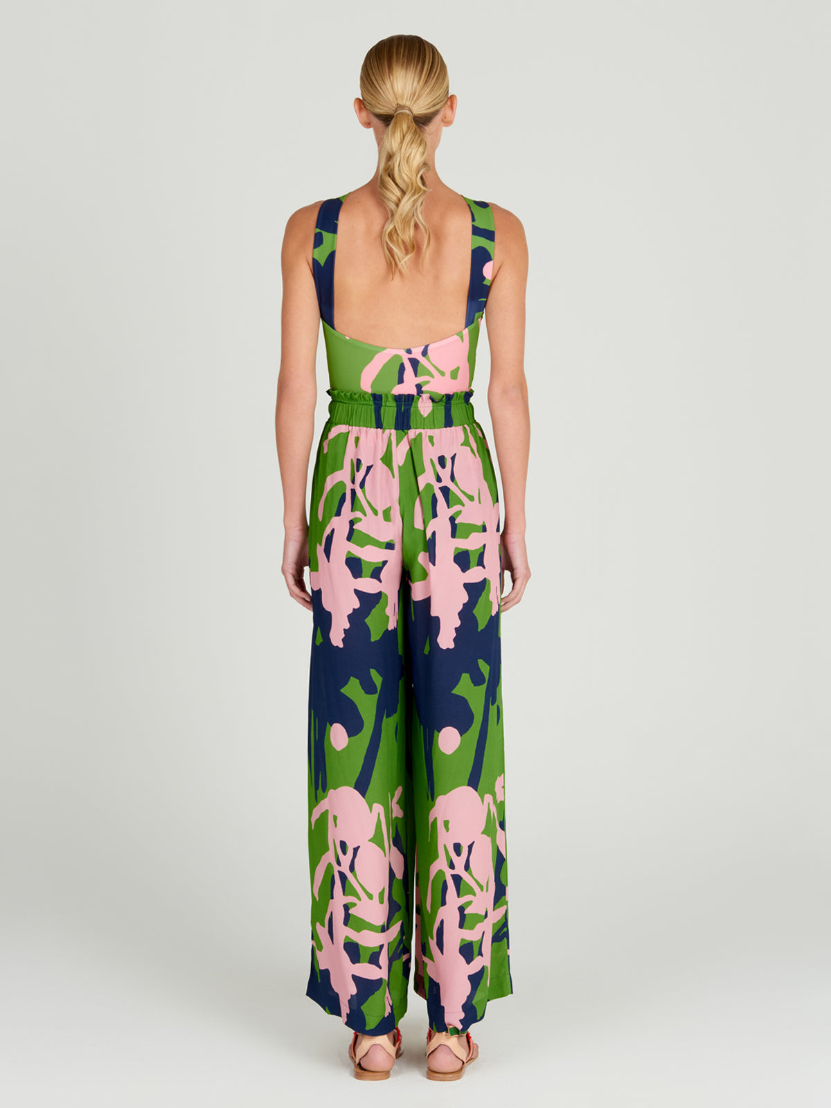 Una mujer con una coleta baja mira hacia otro lado, luciendo el Ovada Pant Verdi Pink: un mono sin mangas con pantalones anchos de cintura alta y un estampado floral abstracto en verde intenso, azul marino y rosa. Completa el look con sandalias.